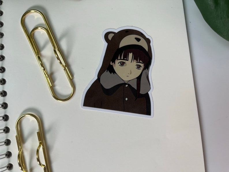 May include: A sticker featuring a cartoon girl with brown hair wearing a brown bear hat and a brown jacket. The girl has a sad expression on her face.