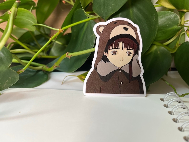 May include: A sticker featuring an anime character wearing a brown bear costume. The character has brown hair and eyes, and is wearing a brown jacket with a zipper. The sticker is on a white background.