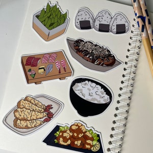 Op de afbeelding: Een set van negen stickers met een Japans eetthema. De stickers bevatten afbeeldingen van sushi, edamame, onigiri, takoyaki, tempura en rijst. De stickers zijn gedrukt op wit papier en hebben een glanzende afwerking.