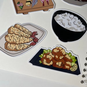Op de afbeelding: Een set van vier stickers met illustraties van Japans eten. De stickers zijn wit met zwarte contouren en tonen een kom rijst, gebakken garnalen, gebakken kip en een bord sushi.