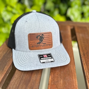 May include: A gray and black trucker hat with a brown leather patch featuring a hockey player silhouette and the text "HEARTWOOD".