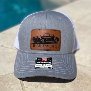 May include: Gray and white trucker hat with a brown leather patch featuring a black silhouette of a vintage car and the text "HEARTWOOD". The hat also has a black and red label with the text "RICHARDSON. ORIGINAL 112 TRUCKER ADJUSTABLE".