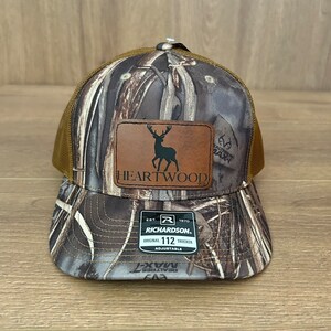 May include: A brown and camouflage trucker hat with a brown leather patch that says "HEARTWOOD" and features a silhouette of a deer. The hat also has a black label that says "RICHARDSON. ORIGINAL 112 TRUCKER ADJUSTABLE".