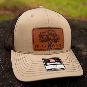 May include: A tan and brown trucker hat with a leather patch featuring a tree design and the text "HEARTWOOD." The hat has a mesh back and a curved brim. A tag reads "RICHARDSON 112 TRUCKER."