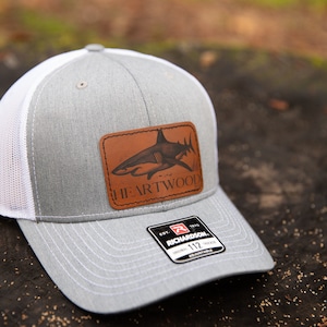 May include: Gray and white trucker hat with a brown leather patch featuring a shark illustration and the text "HEARTWOOD". The hat has a black adjustable strap with a white label that reads "EST. 1970 RICHARDSON 112 ADJUSTABLE".