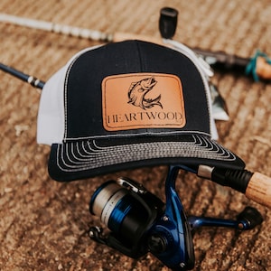 May include: A navy blue and white trucker hat with a brown leather patch featuring a fish graphic and the text "HEARTWOOD 9:13". Fishing rod and blue fishing reel are in the background.