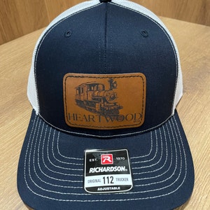 May include: A black and white trucker hat with a brown leather patch featuring a vintage train and the text "HEARTWOOD 9111". The hat has a white mesh back and a black front with a white stitched border. The hat also has a black label with the text "EST. 1970 RICHARDSON. ORIGINAL 112 TRUCKER ADJUSTABLE".