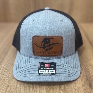 May include: Gray and black trucker hat with a curved brim. The hat features a brown leather patch with a skateboarder graphic and the word "HEARTWOOD". The hat has a Richardson tag.