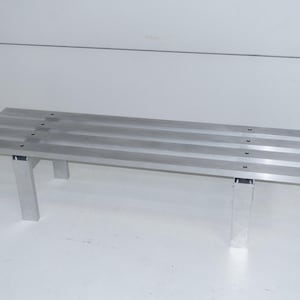 Könnte beinhalten: Eine silberne Metallbank mit vier Latten und zwei Beinen auf jeder Seite. Die Bank ist aus Aluminium und hat ein schlichtes, modernes Design.