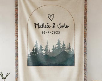 May include: A cream-colored woven throw blanket with a forest scene and the names "Michele & John" and the date "10-7-2025" printed on it. The blanket has a fringed edge and a decorative arch design.