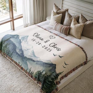 May include: A cream-colored woven throw blanket featuring a mountain landscape design in shades of blue and green. The blanket has the names "Olivia & Chris" and the date "10-28-2025" printed on it, along with a heart and flying birds.