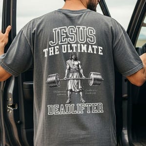 May include: A gray t-shirt with the text "JESUS THE ULTIMATE DEADLIFTER" in white. The graphic depicts Jesus lifting a barbell. The person in the image is wearing a brown cap and dark jeans.