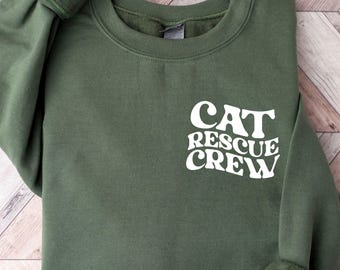 May include: An olive green sweatshirt with the text "CAT RESCUE CREW" in white. The sweatshirt has long sleeves with cuffed wrists and a crew neck. The garment is folded neatly on a light wood-grain surface.