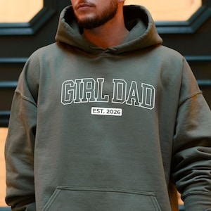 May include: Olive green hoodie with the text "GIRL DAD" in white block letters, with "EST. 2026" below. The hoodie has a front pocket and a drawstring hood.