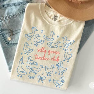 Puede incluir: Camiseta de color marfil con una ilustraci&oacute;n de gansos azules y el texto rosa "silly goose teacher club". La camiseta se muestra con vaqueros azul claro y un sombrero beige. Tambi&eacute;n es visible un collar plateado.