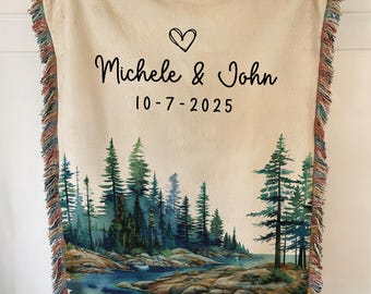 May include: A beige woven throw blanket with a forest scene and the names "Michele & John" and the date "10-7-2025" printed in black. The blanket has a fringe border with shades of brown, red, and blue.