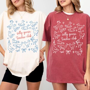 Puede incluir: Dos camisetas holgadas, una crema y otra roja, con un dise&ntilde;o caprichoso. Las camisetas muestran un patr&oacute;n de gansos de dibujos animados, corazones y lazos, con el texto "silly goose teacher club" en una fuente l&uacute;dica. Las camisetas son informales y c&oacute;modas.