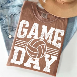 May include: A brown t-shirt with the words "GAME DAY" in large, distressed white letters, along with a volleyball graphic. The shirt is folded on top of a pair of blue jeans and a tan hat.