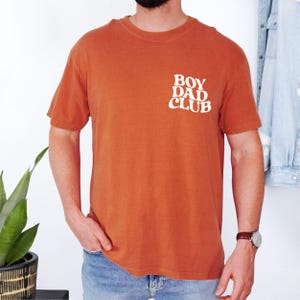 Op de afbeelding: Verbrand oranje t-shirt met de tekst "BOY DAD CLUB" in het wit op de linkerbovenborst. Het shirt heeft korte mouwen en een ronde hals. Het model draagt een blauwe spijkerbroek en een horloge.