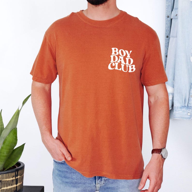 Op de afbeelding: Verbrand oranje t-shirt met de tekst "BOY DAD CLUB" in het wit op de linkerbovenborst. Het shirt heeft korte mouwen en een ronde hals. Het model draagt een blauwe spijkerbroek en een horloge.