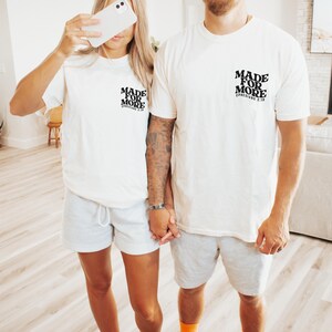 May include: Two people wearing matching white t-shirts and gray shorts. The shirts have the text "MADE FOR MORE" and "EPHESIANS 2:10" printed on the upper left chest. One person is holding a white phone. The other person has tattoos on their arm.