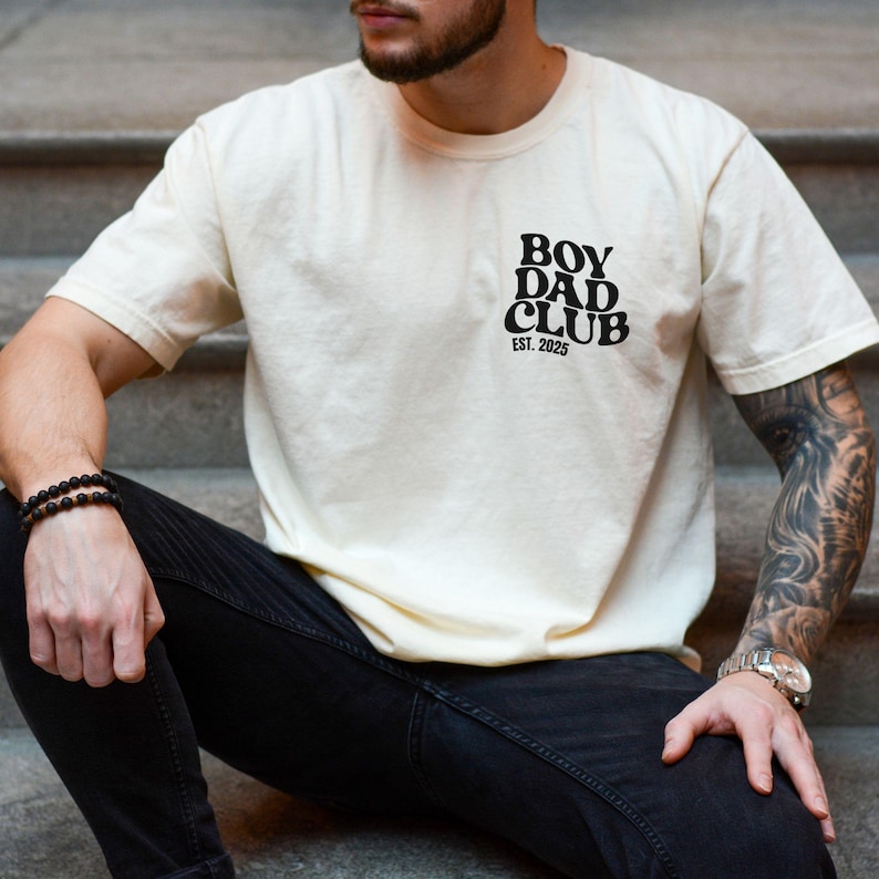 May include: Cream-colored t-shirt with the text "BOY DAD CLUB EST. 2025" printed in black on the upper left chest. The shirt has short sleeves and a relaxed fit. The person is wearing black jeans and a bracelet.