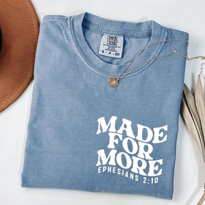 May include: A blue Comfort Colors t-shirt with the text "MADE FOR MORE EPHESIANS 2:10" in white. A gold necklace with a pendant is draped across the shirt. The shirt is folded, and a brown hat is visible in the background.