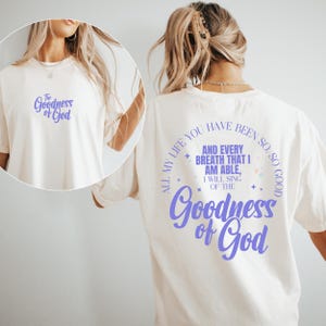 May include: Cream-colored t-shirt with the text "The Goodness of God" in a purple script on the front and back. The back of the shirt features a circular design with the phrase "All my life you have been so good" and more text.
