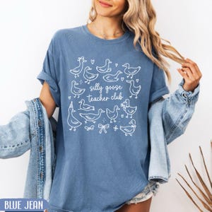 Puede incluir: Camiseta azul vaquero con ilustraciones blancas de gansos y el texto "silly goose teacher club". La camiseta tiene mangas enrolladas y un corte relajado. El dise&ntilde;o incluye corazones, lazos y sombreros de fiesta.