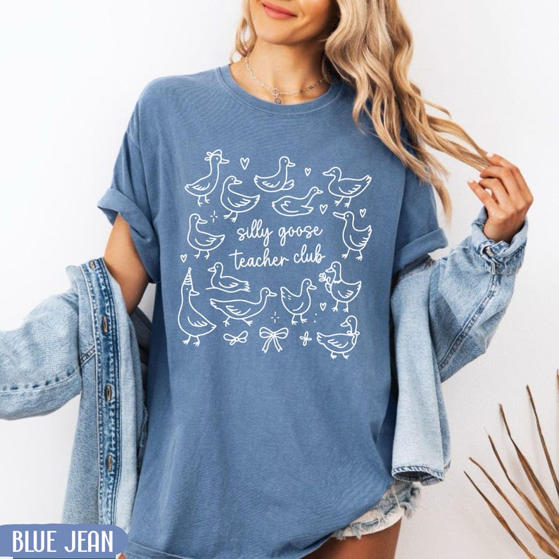 Puede incluir: Camiseta azul vaquero con ilustraciones blancas de gansos y el texto "silly goose teacher club". La camiseta tiene mangas enrolladas y un corte relajado. El dise&ntilde;o incluye corazones, lazos y sombreros de fiesta.