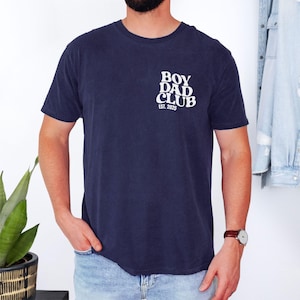 May include: Navy blue t-shirt with the white text "BOY DAD CLUB EST. 2025" on the upper left chest. The shirt is short-sleeved and has a crew neck. A person is wearing the shirt, with their hand in their pocket.