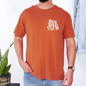 May include: Burnt orange short-sleeve t-shirt with the text "BOY DAD CLUB EST. 2025" printed in white on the upper left chest. The shirt is made of a soft, comfortable material.