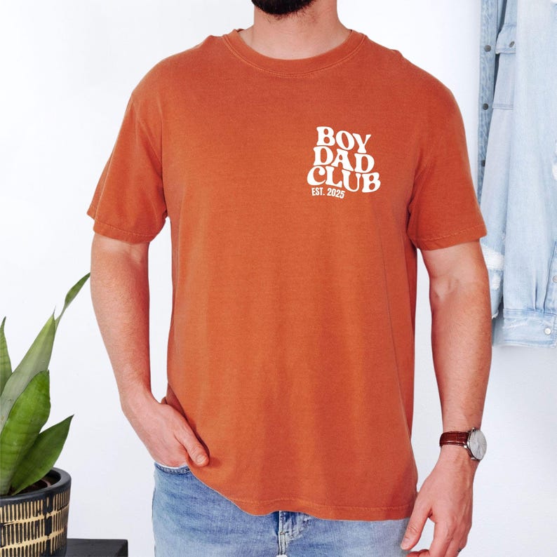 May include: Burnt orange short-sleeve t-shirt with the text "BOY DAD CLUB EST. 2025" printed in white on the upper left chest. The shirt is made of a soft, comfortable material.