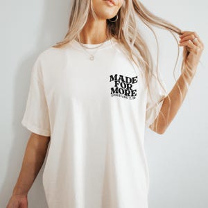 May include: Cream-colored t-shirt with the text "MADE FOR MORE" and "EPHESIANS 2:10" printed in black. The shirt has short sleeves and a relaxed fit. The text is centered on the upper left chest area.