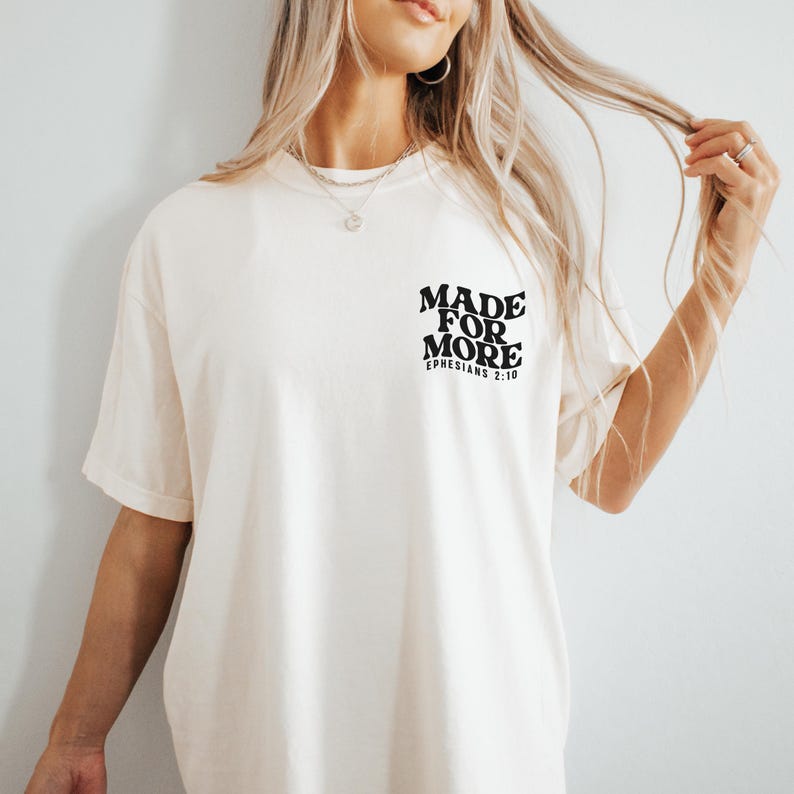 May include: Cream-colored t-shirt with the text "MADE FOR MORE" and "EPHESIANS 2:10" printed in black. The shirt has short sleeves and a relaxed fit. The text is centered on the upper left chest area.