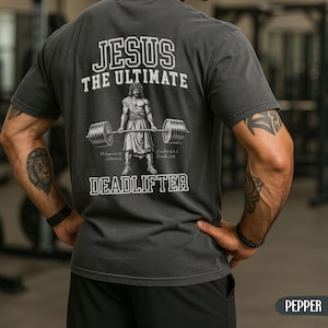 May include: Dark gray t-shirt with the text "JESUS THE ULTIMATE DEADLIFTER" in white. The graphic depicts a muscular figure lifting a barbell. The shirt is a Pepper color.