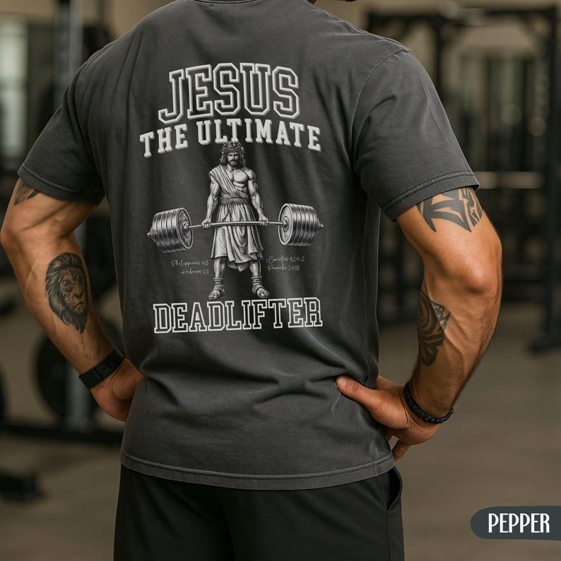 May include: Dark gray t-shirt with the text "JESUS THE ULTIMATE DEADLIFTER" in white. The graphic depicts a muscular figure lifting a barbell. The shirt is a Pepper color.