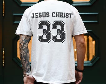 May include: A white short-sleeved t-shirt featuring the text JESUS CHRIST in a black varsity-style font above the large number 33. The number 33 is printed in a distressed black font with a white outline, mimicking a sports jersey design.