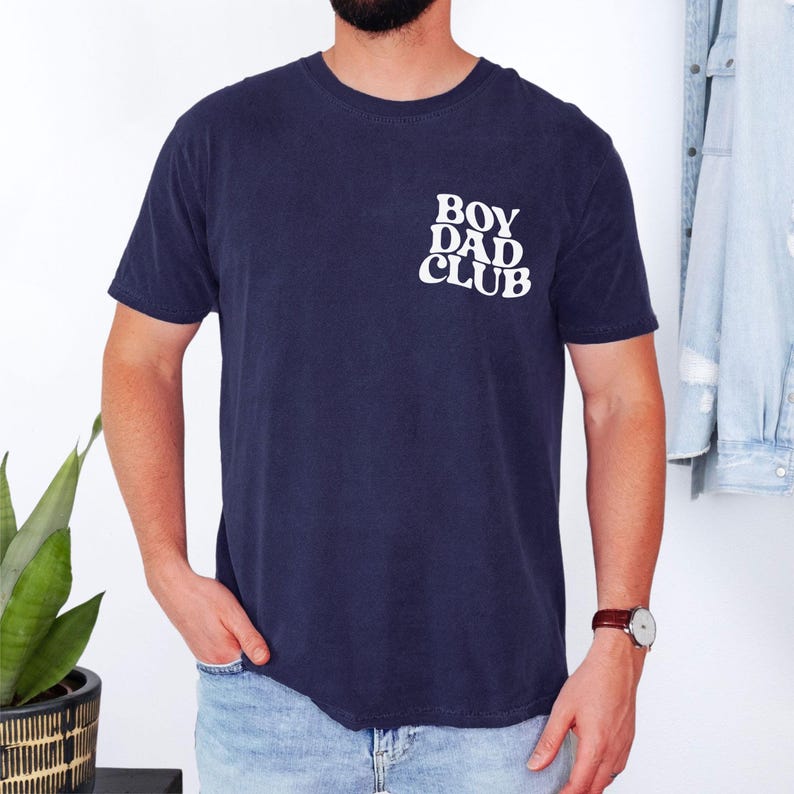 Op de afbeelding: Marineblauw T-shirt met de witte tekst "BOY DAD CLUB" op de borst. Het shirt heeft korte mouwen en een ronde hals. Het model draagt een horloge met een bruine leren band.