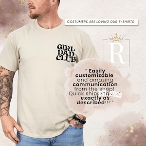 Op de afbeelding: Een beige t-shirt met de woorden "GIRL DAD CLUB" in het zwart gedrukt op de linkerbovenborst. Het shirt heeft ook een tekstballon met tekst. De bovenkant van de afbeelding luidt "COSTUMERS ARE LOVING OUR T-SHIRTS".