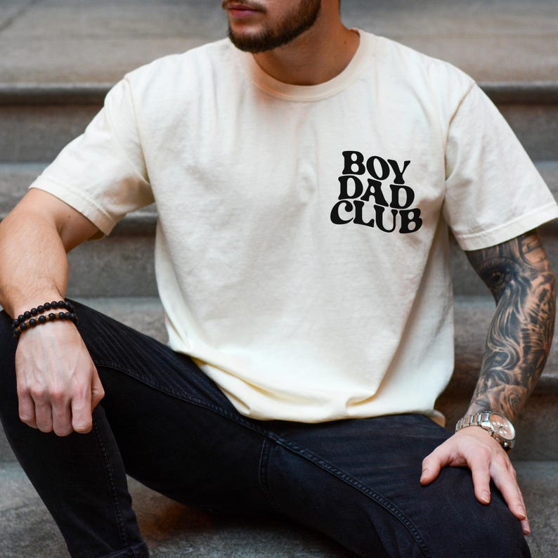 Op de afbeelding: Cr&egrave;mekleurig T-shirt met de zwarte tekst "BOY DAD CLUB". Het shirt heeft korte mouwen en lijkt van een zacht materiaal te zijn. De persoon op de foto draagt een zwarte jeans en een horloge.