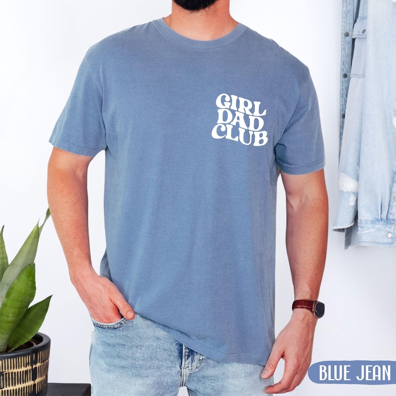 Op de afbeelding: Blauw T-shirt met korte mouwen met de witte tekst "GIRL DAD CLUB" op de linkerbovenborst. Het shirt is effen blauw van kleur en gemaakt van een zacht materiaal. De woorden "BLUE JEAN" staan in een blauwe rechthoek rechtsonder.
