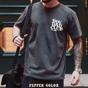 May include: A dark gray t-shirt with the text "BOY DAD CLUB EST. 2025" in white. The shirt is a pepper color and has a vintage look. The image shows the shirt on a person.
