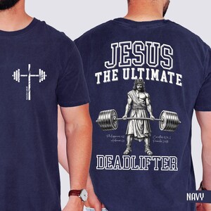 May include: Navy blue t-shirt with a white barbell cross graphic on the front. The back features a graphic of Jesus lifting a barbell with the text "JESUS THE ULTIMATE DEADLIFTER". Includes Bible verses.