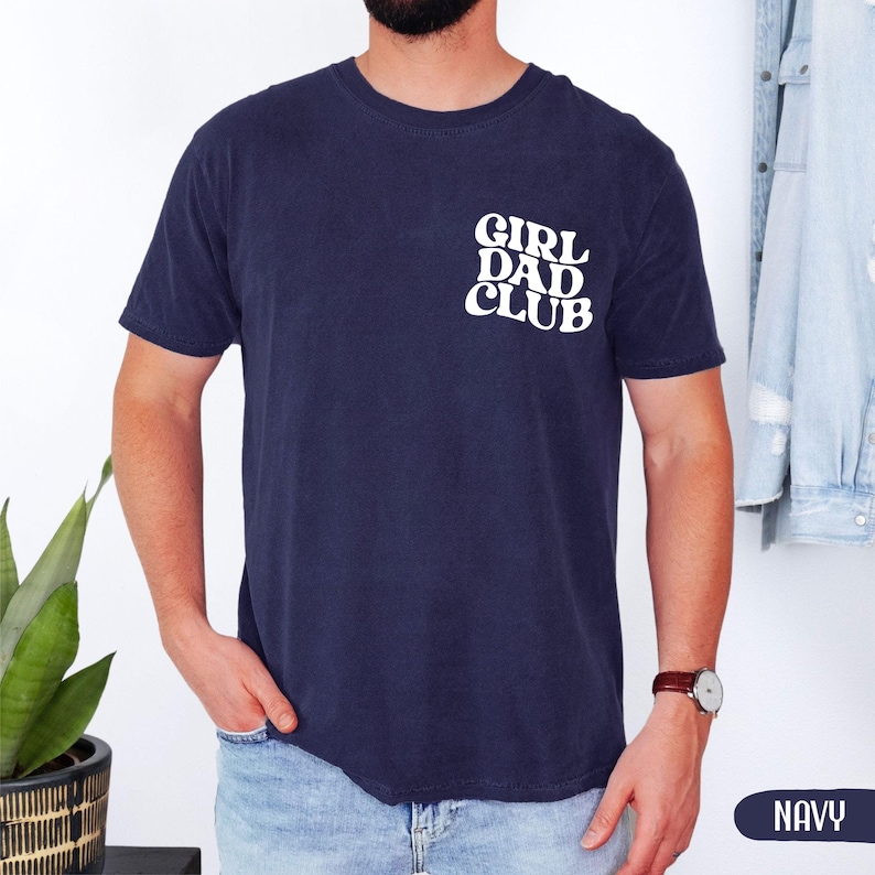 Op de afbeelding: Marineblauw T-shirt met de witte tekst "GIRL DAD CLUB" op de linkerborst. Het shirt met korte mouwen is effen van kleur. Het woord "NAVY" wordt in een witte rechthoek rechtsonder weergegeven.