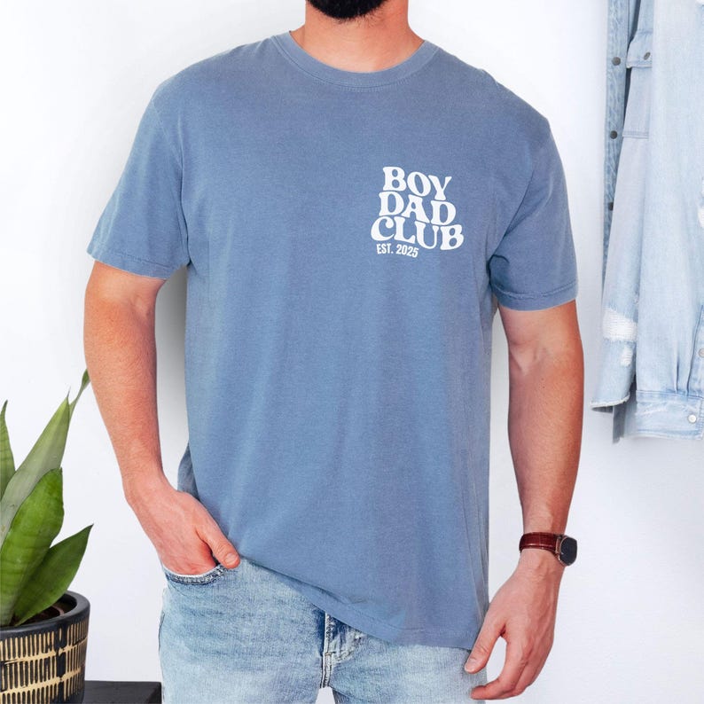 May include: A dusty blue t-shirt with the text "BOY DAD CLUB EST. 2025" printed in white on the upper left chest. The shirt has short sleeves and a crew neck. The person is wearing blue jeans and a brown watch.