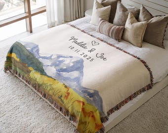 May include: A cream-colored throw blanket with a watercolor mountain scene in blue, green, and yellow. The blanket has a fringed edge and features the names "Maddie & Joe" and the date "10.11.2025" in black script.