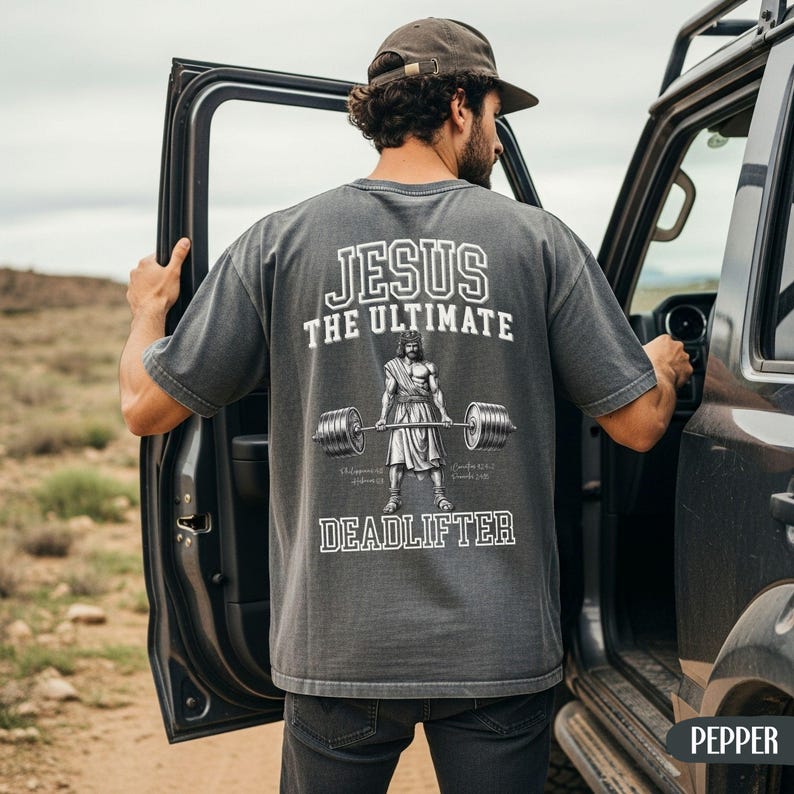 May include: A gray t-shirt with the text "JESUS THE ULTIMATE DEADLIFTER" in white. The graphic depicts Jesus lifting a barbell. The person in the image is wearing a brown cap and dark jeans.