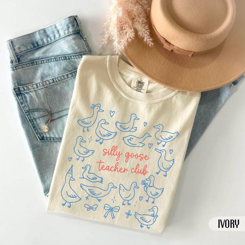 Puede incluir: Camiseta de color marfil con una ilustraci&oacute;n de gansos azules y el texto rosa "silly goose teacher club". La camiseta se muestra con vaqueros azul claro y un sombrero beige. Tambi&eacute;n es visible un collar plateado.