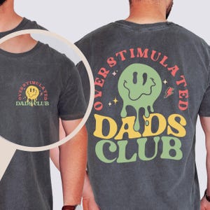 May include: Dark gray t-shirt with the text "OVERSTIMULATED DADS CLUB" in a retro font. The design includes a green melting smiley face, stars, and a lightning bolt. The same design is on the front and back of the shirt.